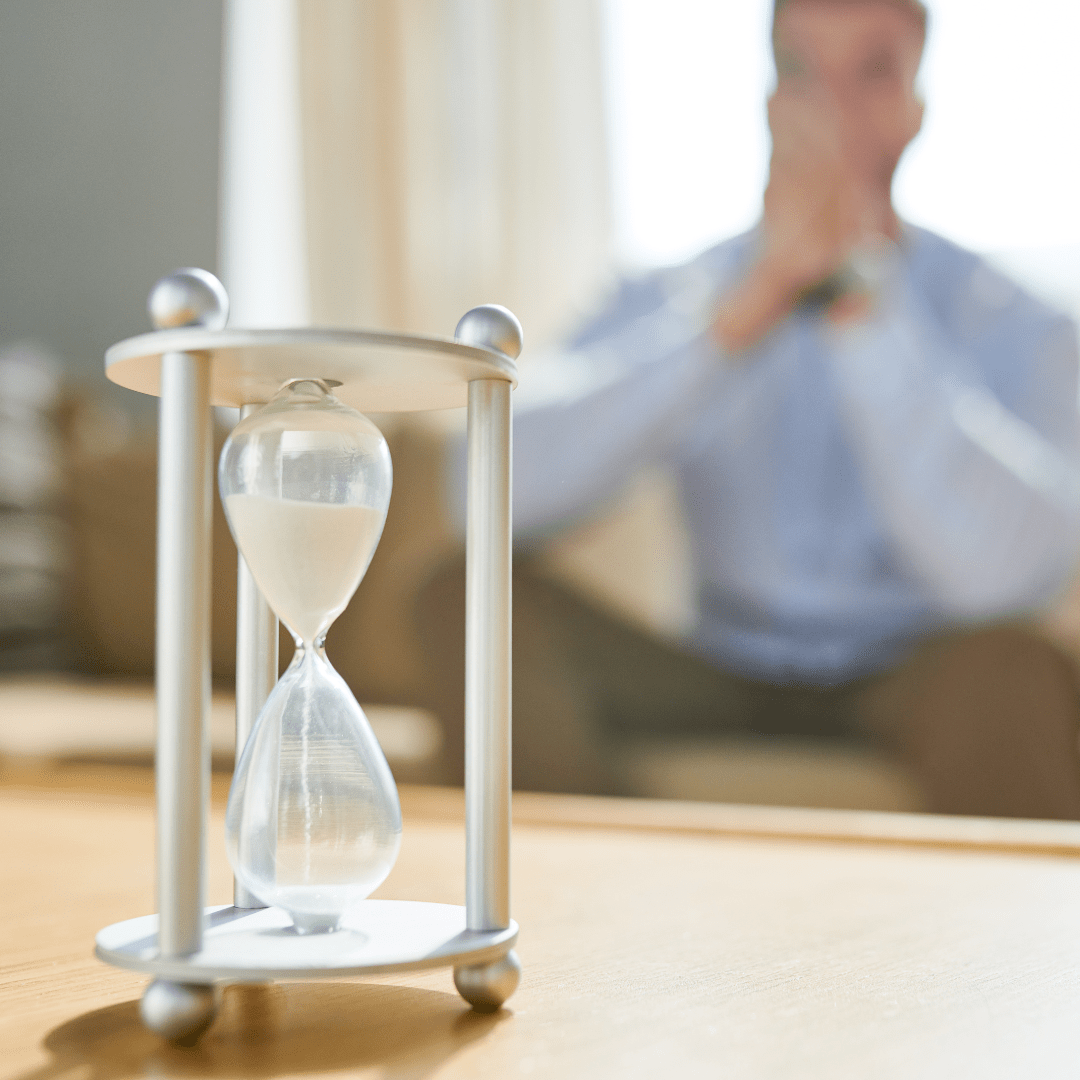 Hourglass on a table with a blurred person in the background, symbolizing the waiting period before a Business Security Check-In