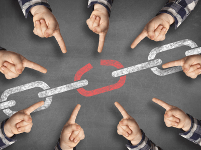 Multiple hands point toward a broken red link in a chalk-drawn chain on a blackboard, symbolizing a weak point in a system or process.